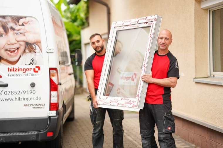 Zwei Monteure in roten Hilzinger-Shirts tragen ein Fenster neben dem Firmenbus